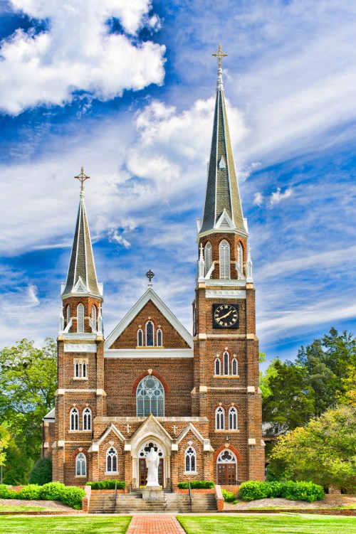 A tall, brick church with two spires, a clock, and lush greenery under a blue sky with clouds.