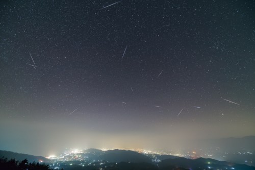 A starry night sky filled with shooting stars over a cityscape illuminated by lights below.