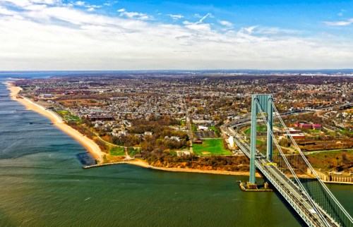 Aerial view of a bridge spanning a river, with a coastal landscape and urban area in the background under a clear sky.