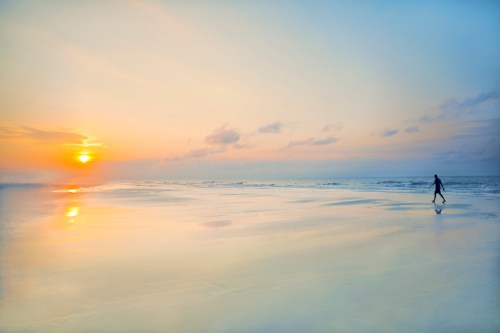 A person walks along a serene beach at sunset, with soft waves and a colorful sky reflecting on the wet sand.