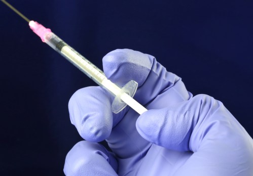 A gloved hand holds a syringe with a pink cap against a dark background.