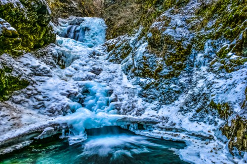 A frozen waterfall cascading down rocky cliffs, surrounded by icy formations and a serene turquoise pool below.