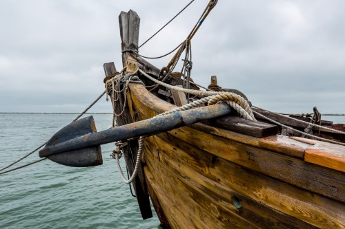 A close-up of a wooden boat's bow with an anchor and ropes, set against a cloudy sky over calm waters.