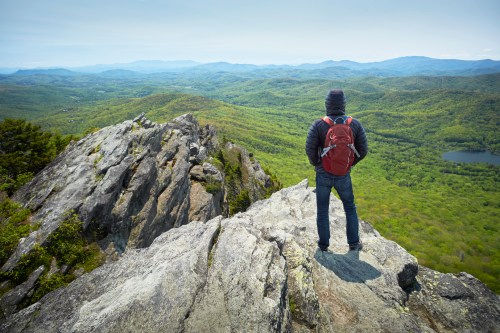 A person stands on a rocky ledge, overlooking a lush green valley and distant mountains under a clear sky.