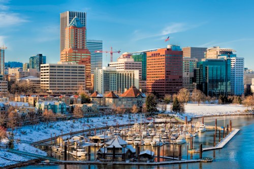 A scenic view of a city skyline with modern buildings, a marina, and snow-covered ground under a clear blue sky.