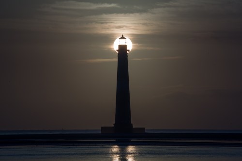 A silhouette of a lighthouse against a sunset, with the sun glowing behind its light.