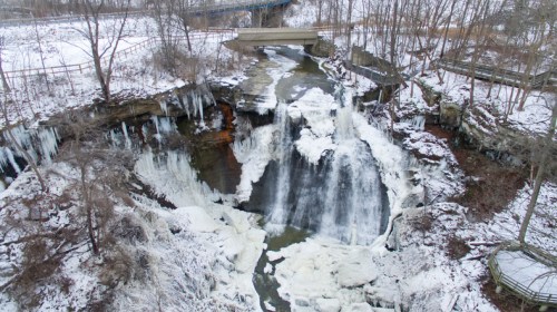 A frozen waterfall surrounded by snow-covered rocks and trees, with a bridge in the background.