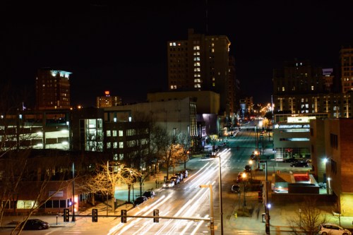 A city street at night, illuminated by streetlights and car headlights, with buildings lining the road.