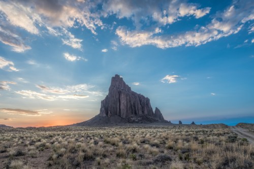 A dramatic rock formation rises against a colorful sunset sky, surrounded by open grassland and distant mountains.