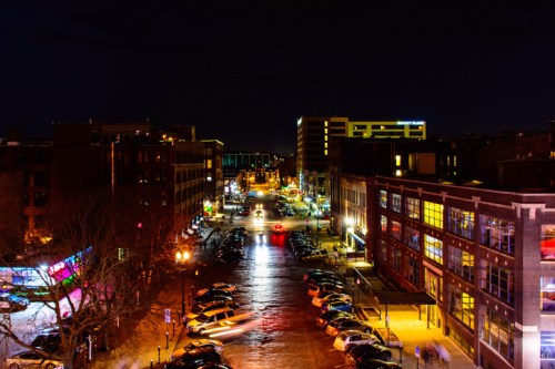A city street at night, illuminated by streetlights and car headlights, with buildings lining both sides.