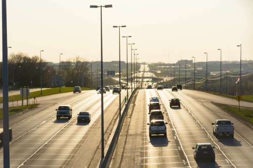 A sunlit highway with cars traveling in both directions, lined with streetlights and greenery on the sides.