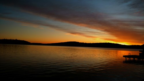 Sunset over a calm lake, with vibrant orange and purple hues reflecting on the water and silhouettes of distant hills.