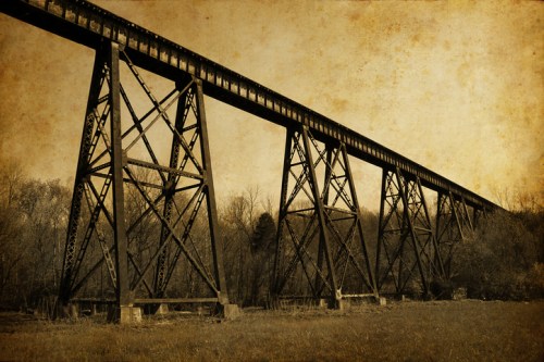 A vintage-style image of a tall, metal railway bridge spanning over a grassy area, surrounded by trees.