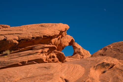 A natural rock formation with an arch, set against a clear blue sky and a small crescent moon in the background.