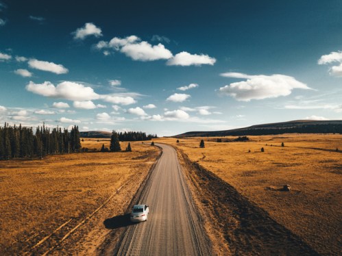 A car drives along a winding dirt road through a golden field under a blue sky with fluffy clouds.