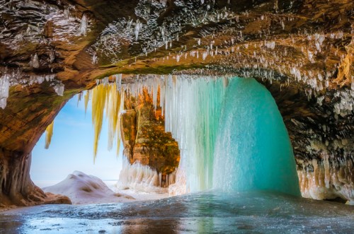A stunning ice cave with icicles hanging from the ceiling and a turquoise waterfall flowing into a frozen landscape.