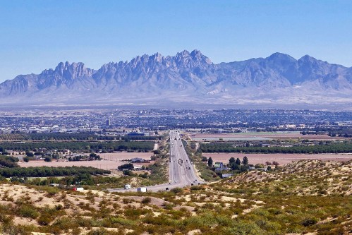 A wide view of a highway leading towards distant mountains, with fields and vegetation in the foreground.