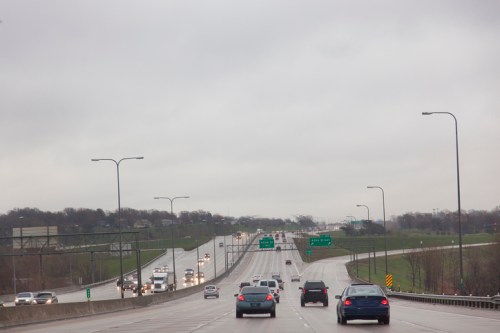 A view of a highway on a cloudy day, with cars traveling in both directions and green highway signs visible.