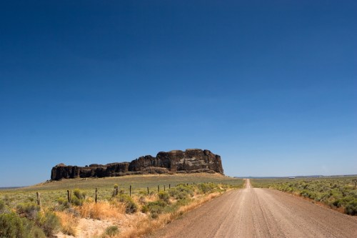 A dirt road leads to a large rock formation under a clear blue sky, surrounded by green grass and sparse vegetation.