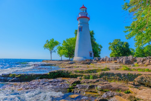A tall lighthouse stands by the shore, surrounded by trees and rocky terrain, with a clear blue sky and water.