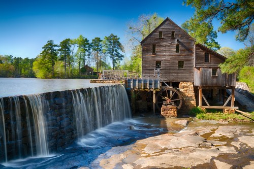 A historic watermill beside a serene lake, featuring a wooden structure and a flowing waterfall. Lush greenery surrounds the scene.