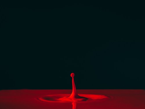 A single red droplet splashes upward against a dark background, creating ripples in a pool of red liquid.