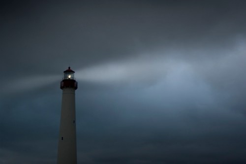 A tall lighthouse stands against a dark, cloudy sky, with its light beam cutting through the gloom.