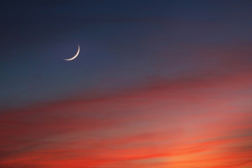 A crescent moon shines in a twilight sky, surrounded by soft hues of red and blue.