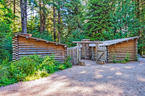 A rustic log cabin structure surrounded by tall trees and greenery, set on a gravel path.
