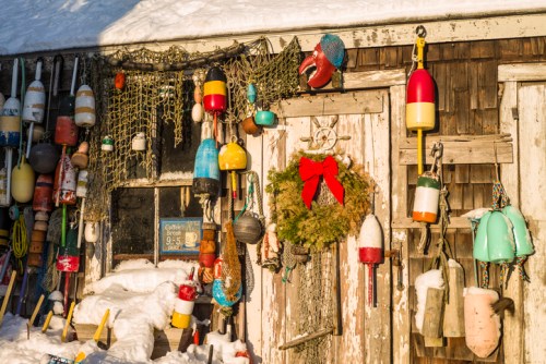 A rustic shed adorned with colorful buoys, fishing gear, and a festive wreath, surrounded by snow.