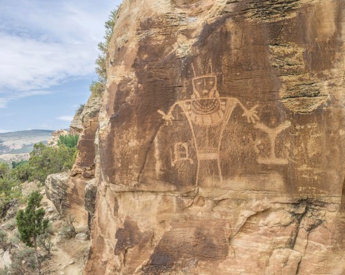 Petroglyph of a figure with outstretched arms carved into a rocky cliff, surrounded by trees and a scenic landscape.