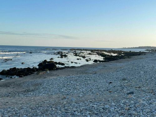 A serene beach scene with smooth pebbles, rocky shoreline, and gentle waves under a clear blue sky.