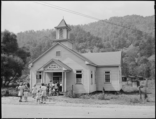 A historic church building with a steeple, surrounded by a group of people outside on a rural road.