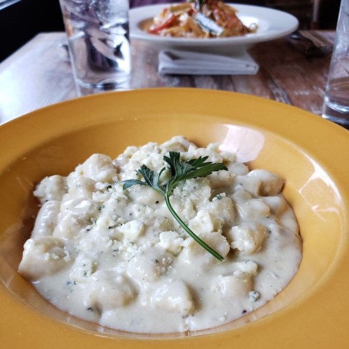 A creamy plate of gnocchi topped with herbs, served in a yellow bowl, with another dish in the background.