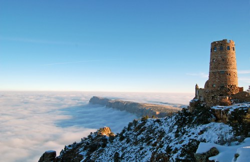 A stone tower stands on a snowy cliff above a sea of clouds, with distant mountains visible under a clear blue sky.