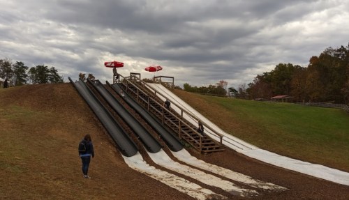 A grassy hill with three sliding lanes, people sliding down, and cloudy skies above.