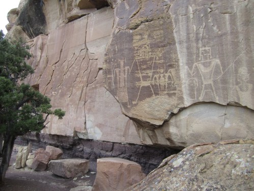 Petroglyphs carved into a large rock face, depicting figures and symbols, surrounded by natural rock formations.