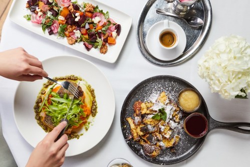A table set with a plate of pasta, a salad, dessert, coffee, and a vase of flowers, with a hand preparing to eat.