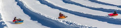 Three people tubing down snowy slopes, surrounded by snow-covered hills and bright blue skies.