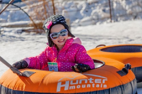 A smiling child in a pink jacket and sunglasses sits in a snow tube at Hunter Mountain, surrounded by snow.