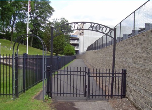 A gated entrance with an archway labeled "Ed Moore," leading to a pathway beside a stone wall and grassy area.