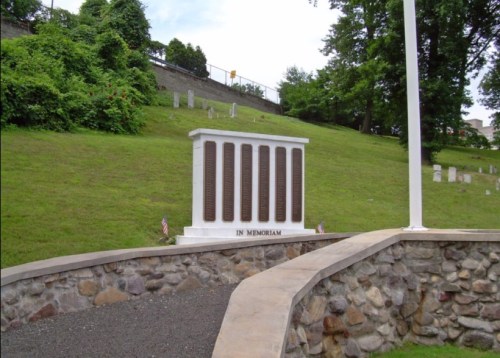 A memorial monument with engraved plaques, surrounded by greenery and a stone pathway. Flags are placed nearby.