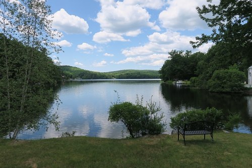 A serene lake view surrounded by greenery, with fluffy clouds reflecting in the water under a blue sky.