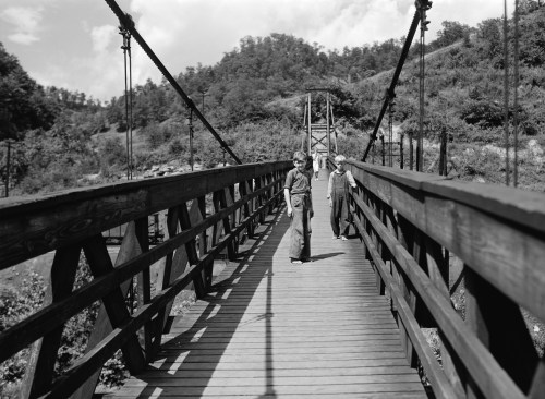 Two children walking on a wooden suspension bridge surrounded by trees and hills in a black and white photograph.