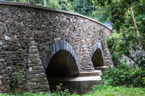 A stone bridge with arched supports spans a small creek, surrounded by lush greenery and trees.