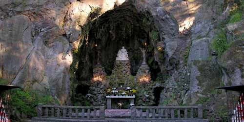 A serene cave altar with a statue, surrounded by candles and flowers, set against rugged stone walls.