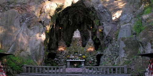 A serene cave altar with a statue, surrounded by candles and flowers, set against rugged stone walls.