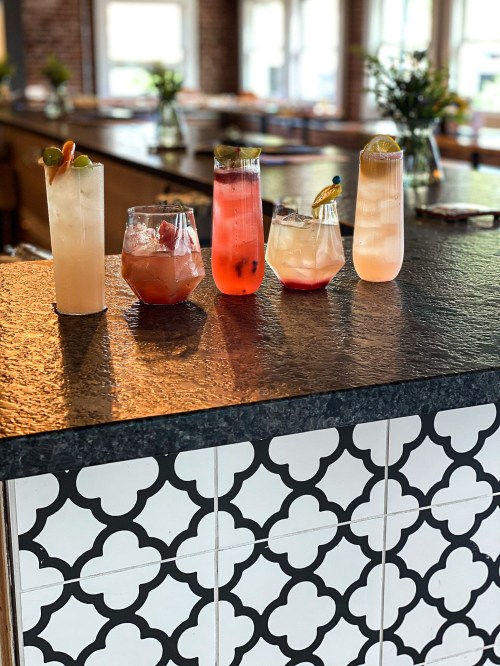 Five colorful cocktails on a bar counter, featuring various garnishes and glass styles, with a patterned tile backdrop.