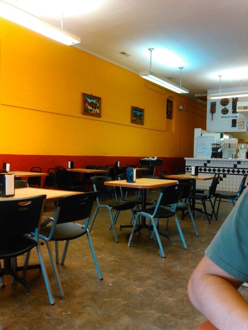 Interior of a casual restaurant with empty tables, colorful walls, and artwork. A person’s arm is partially visible.