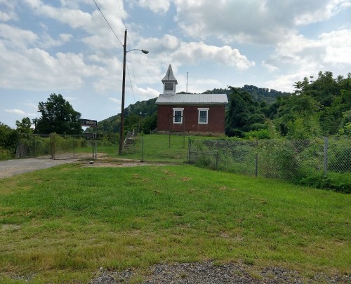 A small brick building with a steeple sits on a grassy hill, surrounded by a fence and trees in the background.